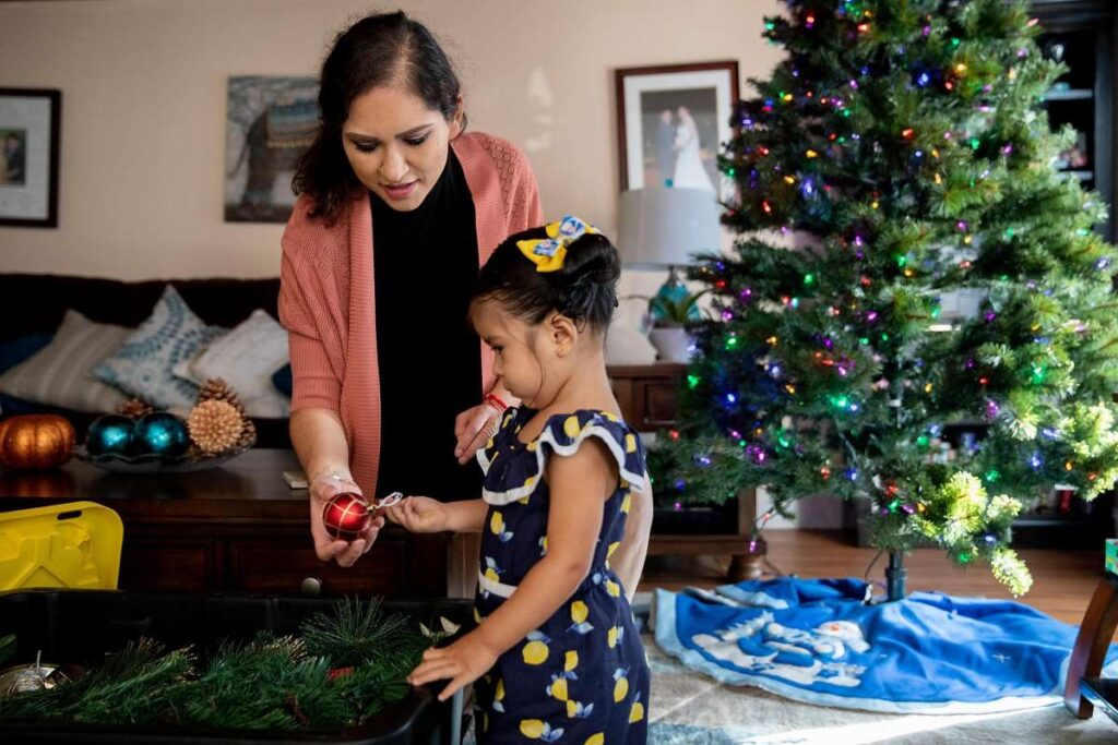 Mother and daughter smile while hanging ornaments on a Christmas tree.