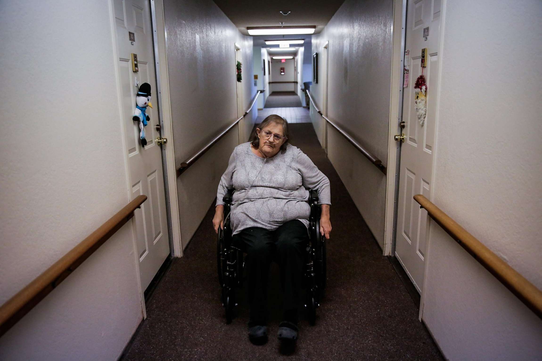 Elderly woman in a wheelchair moving down a hallway.