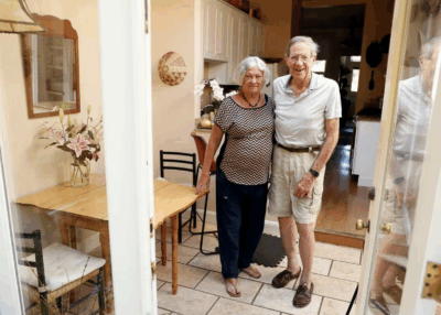 Elderly couple stands together in a warmly lit home.