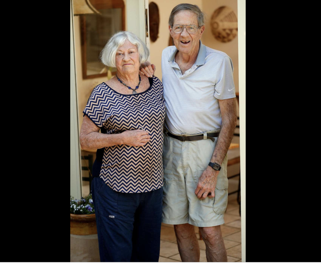 Elderly couple stands together in a doorway, smiling.