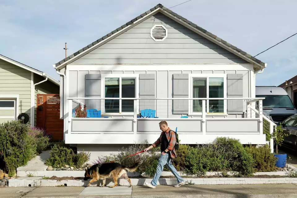 A man walks his dog past a small house—illustrating independence and daily life.