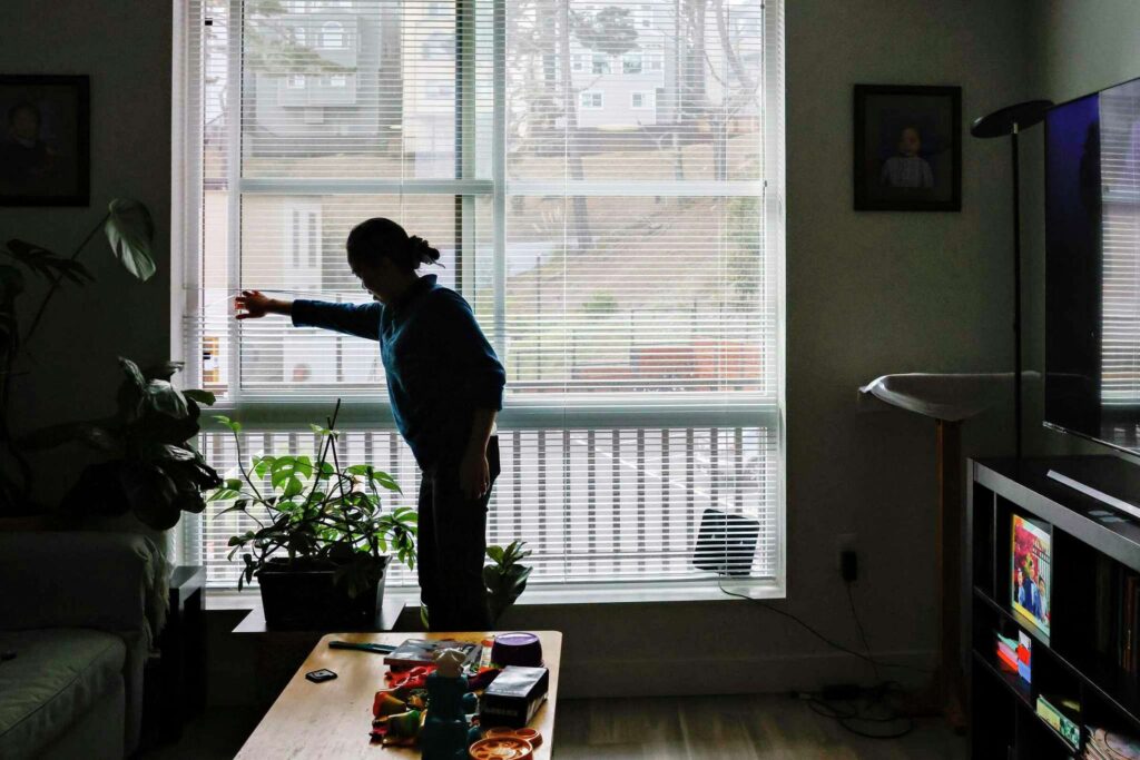 A woman looks at houseplant near a large window—depicting routine, growth, and calm.