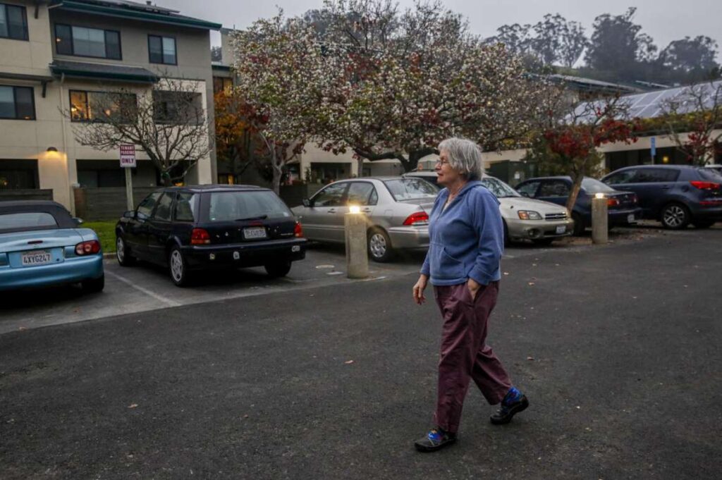 An elderly woman walks through a parking lot with hands in his pockets—conveying daily routine and quiet independence.
