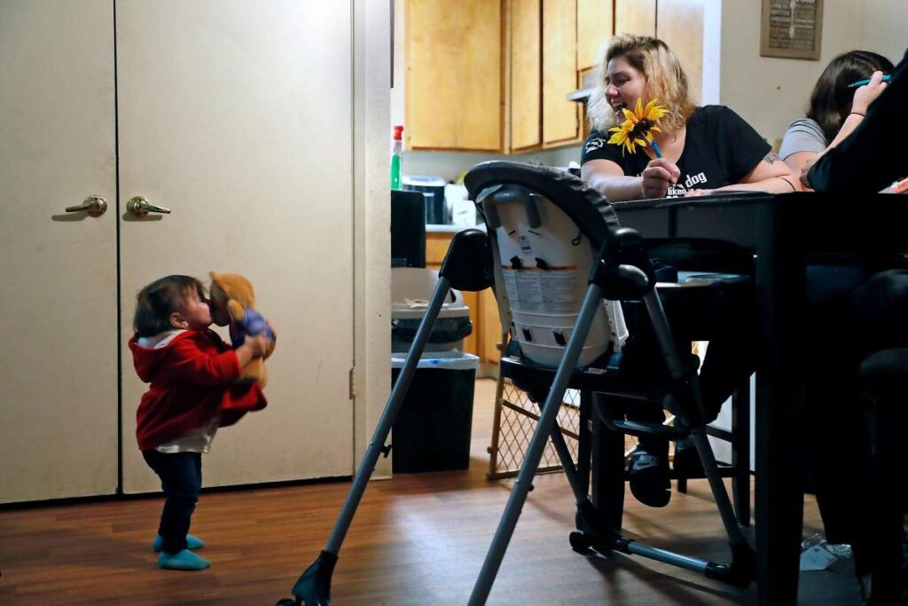 Mother sits at table while smiling at child playing with doll.