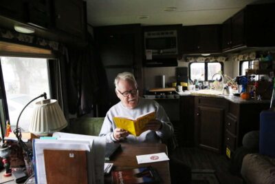 Elderly man reads a book inside his trailer.