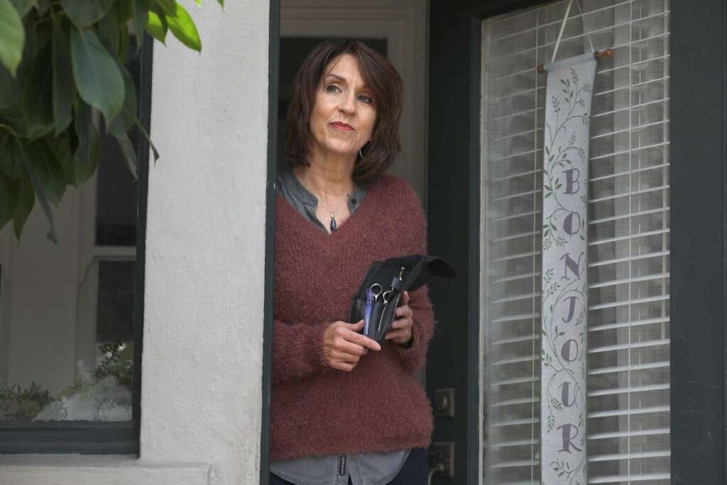 Woman holding haircutting tools, stands on a porch.