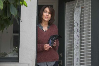 Woman holding haircutting tools, stands on a porch.