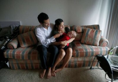 Young couple sitting on a couch, smiling while holding their baby together in a warm, home setting.