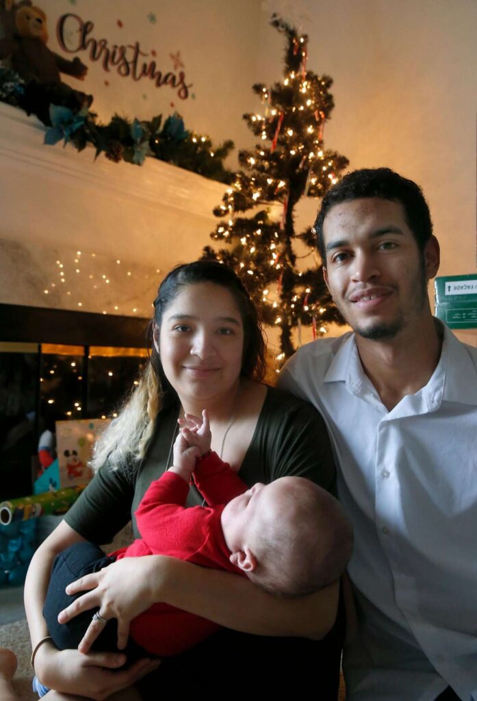 New parents holding their baby and smiling in front of a lit Christmas tree, conveying warmth and family joy during the holidays.
