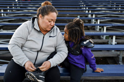 A woman ties a young girl’s shoelaces in stadium seating—showing love and tradition.