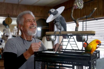 Older man interacts with a parrot perched on a cage.