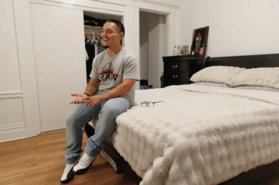 Young man sits on bed smiling.